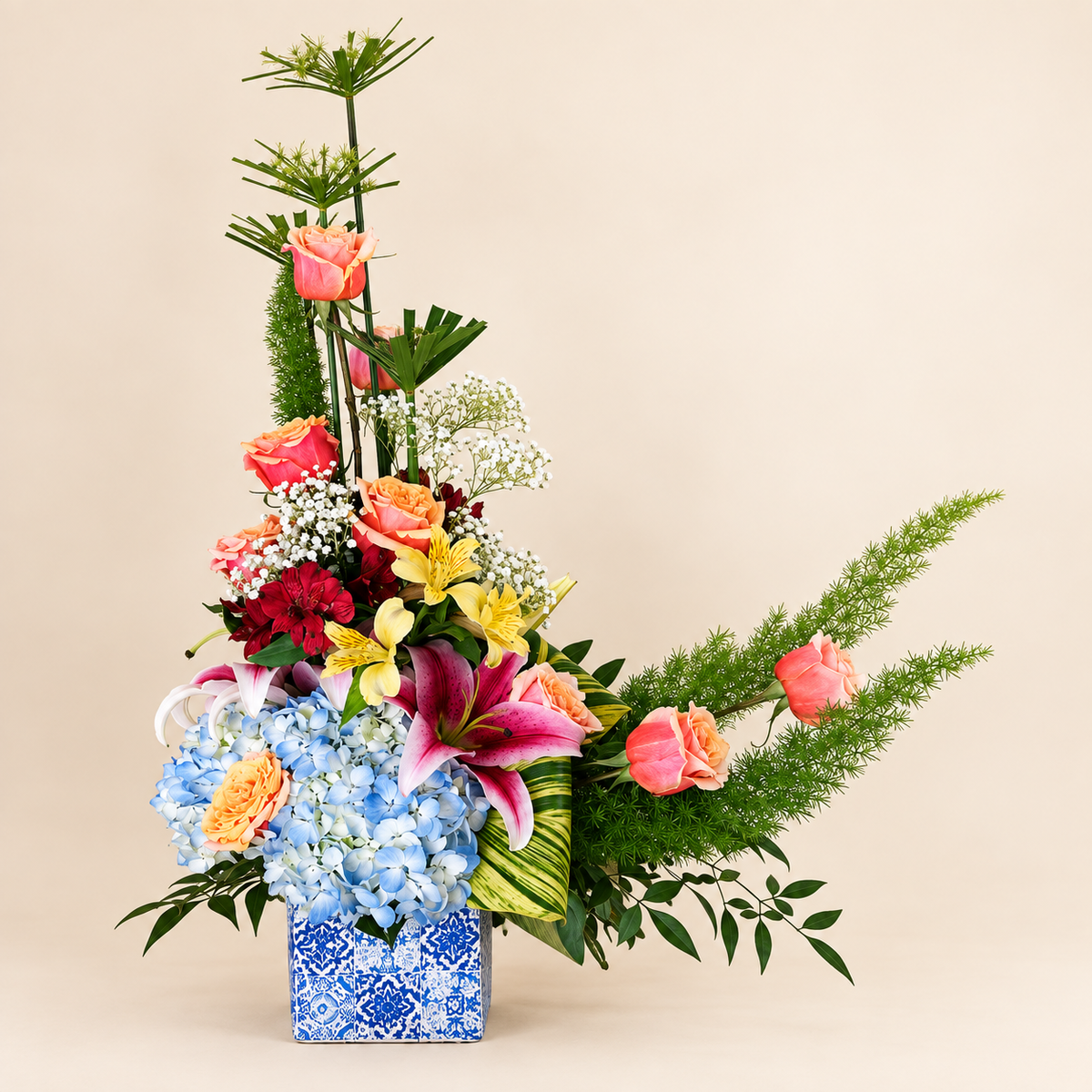 Blue Horizon Garden arrangement with blue hydrangea, coral roses and lilies in a patterned vase on ivory