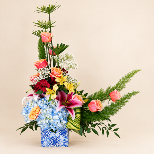 Blue Horizon Garden arrangement with blue hydrangea, coral roses and lilies in a patterned vase on ivory