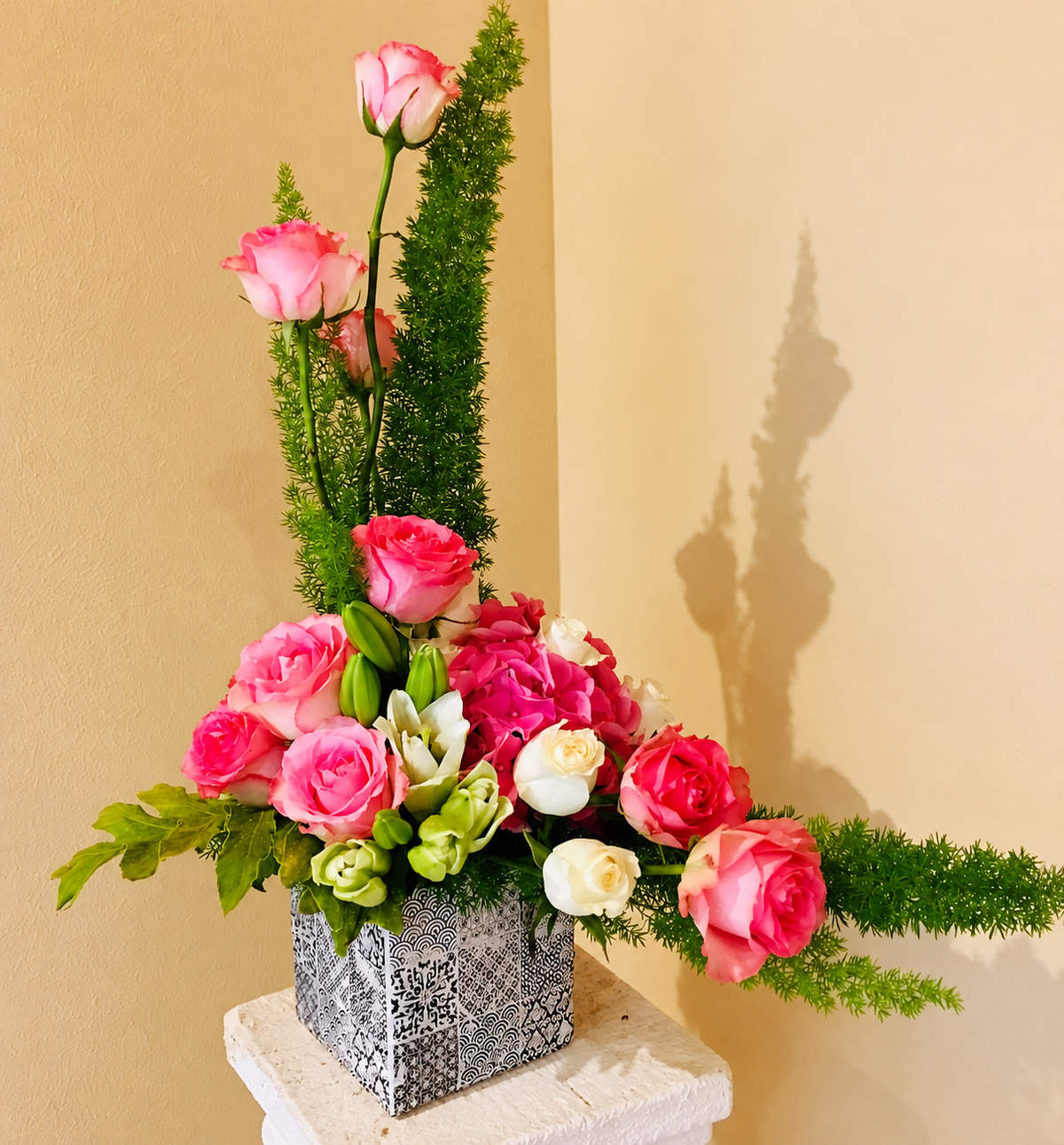 Rose and fern arrangement in its patterned square vase, photographed on a stone pedestal against a warm wall
