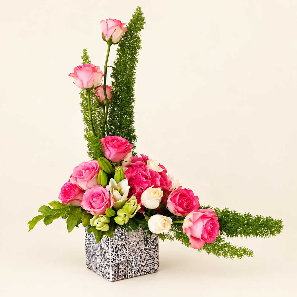 Sculptural pink rose and greenery arrangement in a patterned square vase on an ivory background