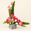 Sculptural pink rose and greenery arrangement in a patterned square vase on an ivory background