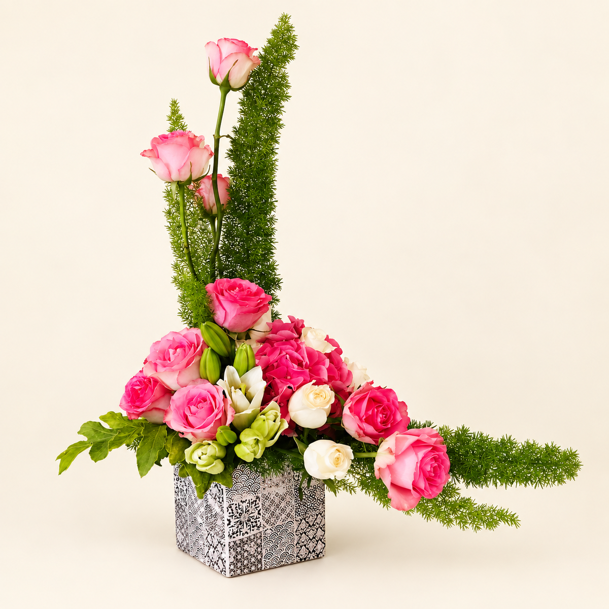 Sculptural pink rose and greenery arrangement in a patterned square vase on an ivory background