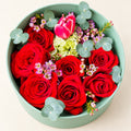 Red roses, a pink tulip, green hydrangea, waxflower and eucalyptus in a seafoam round flower gift box on an ivory background