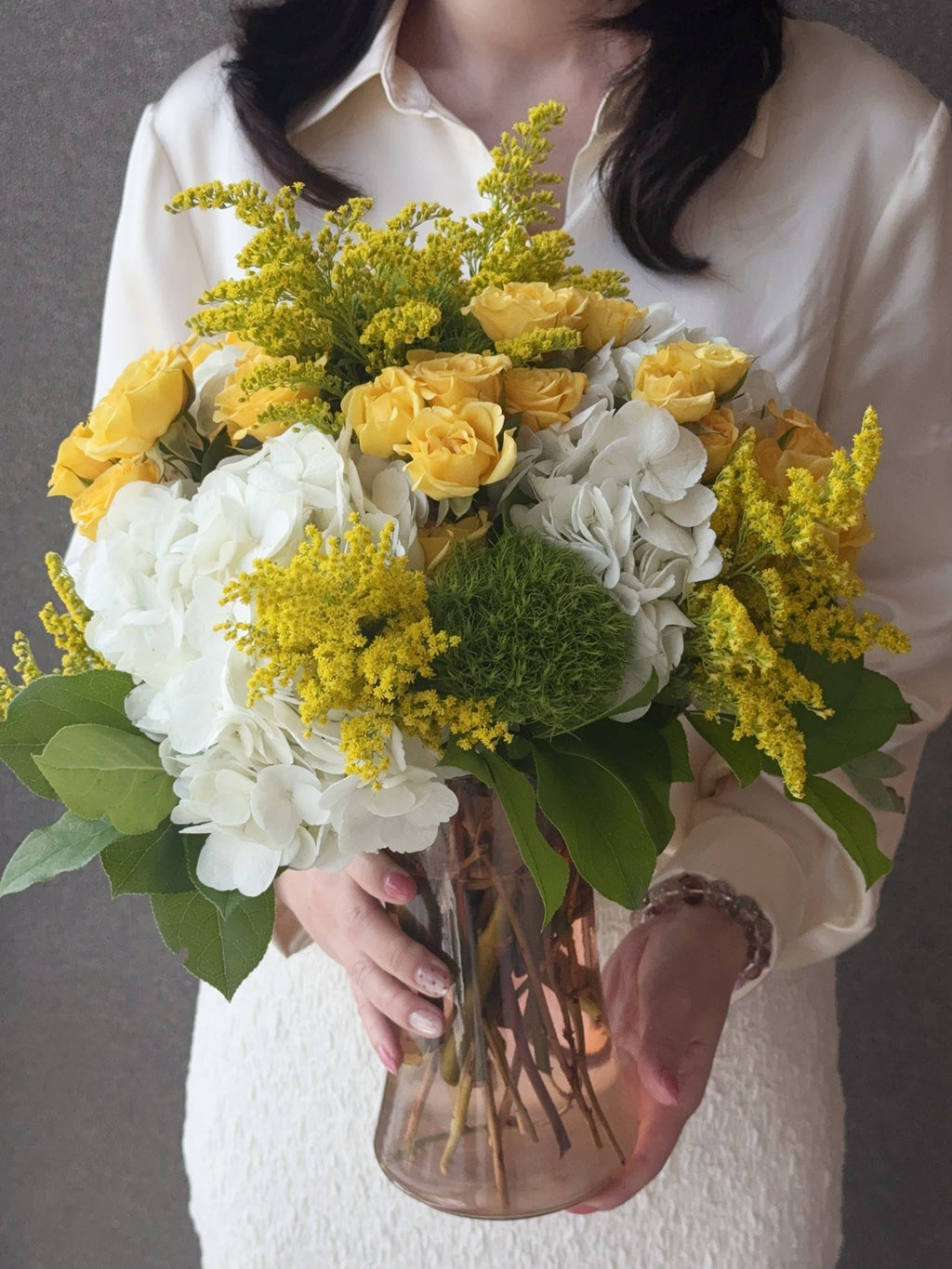 Original photo of a person holding the Sunlit Harmony arrangement with yellow roses, white hydrangeas and greenery in a glass vase